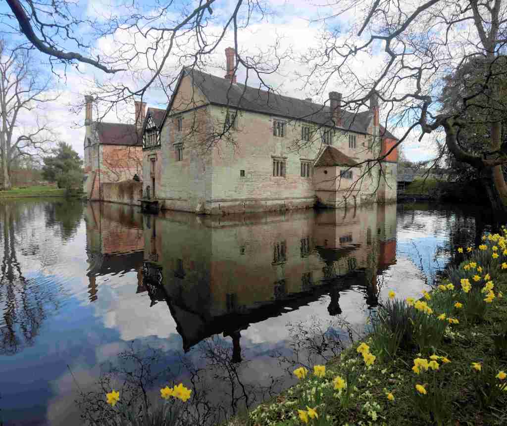 Baddesley Clinton moated manor house in Warwickshire, viewed across the moat with daffodils in the foreground.